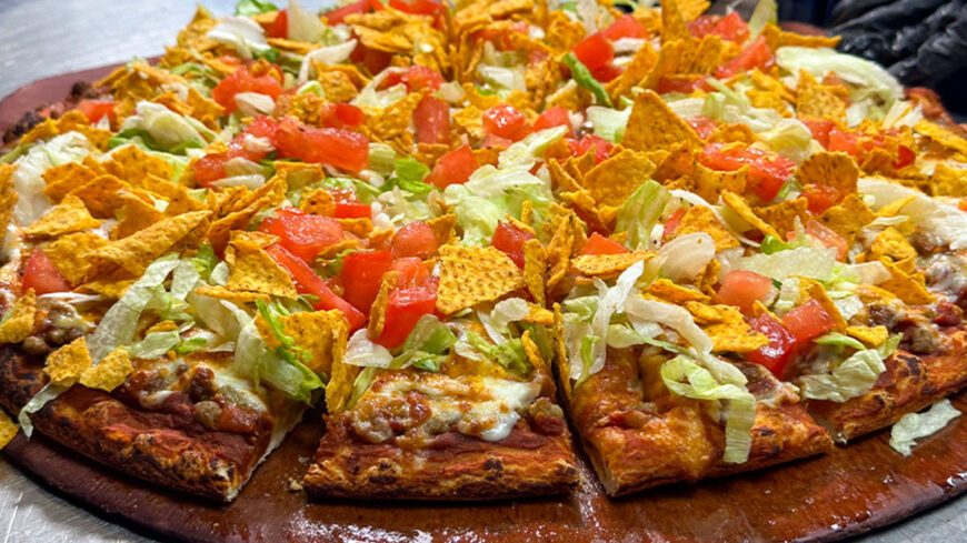 Pizza topped with lettuce, diced tomatoes, seasoned corn chips, beans, meat, and melted cheese on a wooden serving board.