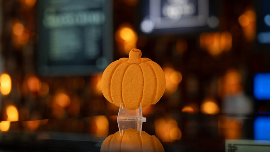 A pumpkin-shaped golden-brown cookie displayed on a clear stand in a shop window, with warm, glowing lights blurred in the background.