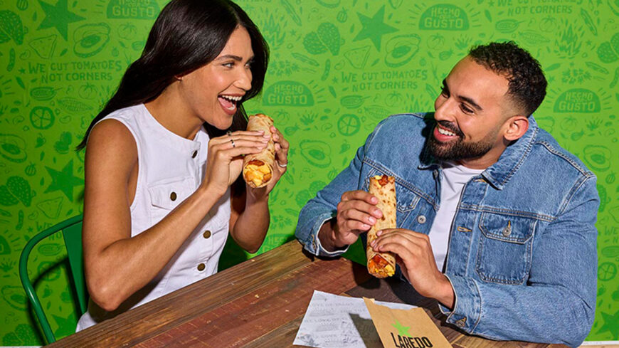 Two people smiling and eating tacos at a table in front of a bright green Laredo Taco Company backdrop.