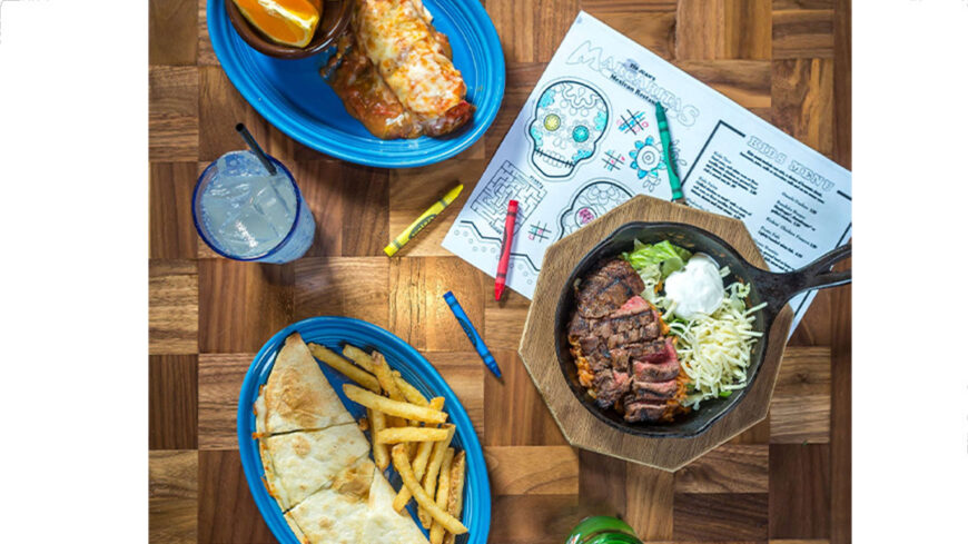 Overhead view of a family-friendly meal on a wooden table, featuring a steak skillet with rice, cheese, and sour cream, a quesadilla with fries, and another entrée, alongside drinks and a children’s coloring activity sheet.
