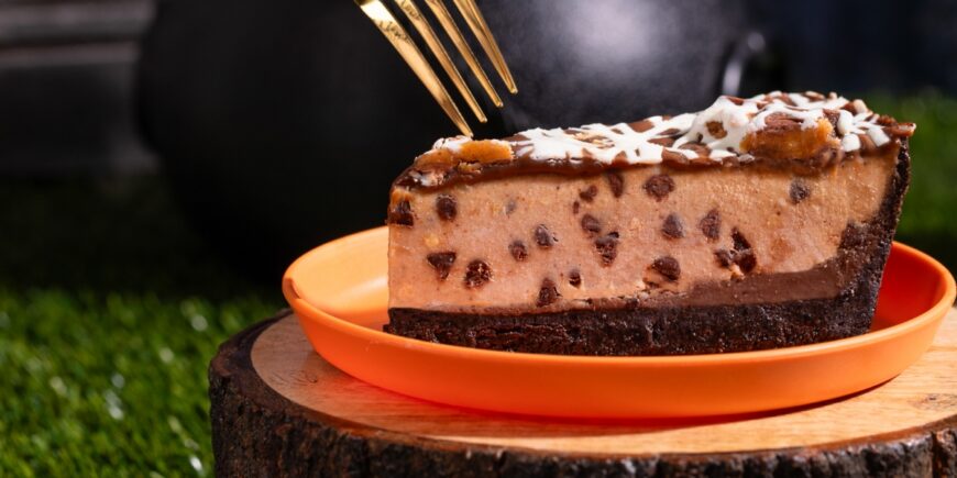 Slice of Reese’s peanut butter pie featuring creamy peanut butter filling with chocolate pieces, a chocolate cookie crust, and chocolate and white icing drizzles, served on an orange plate with a fork above it.