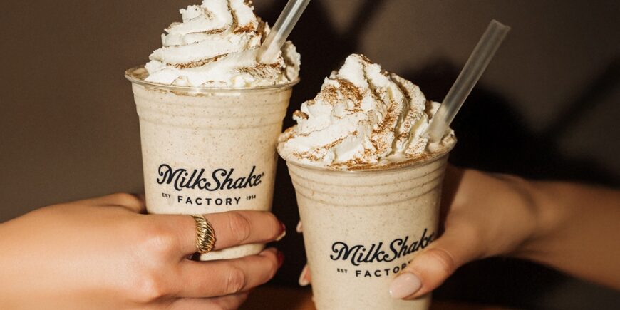 Two hands hold MilkShake Factory milkshakes topped with generous swirls of whipped cream and cinnamon in branded cups.