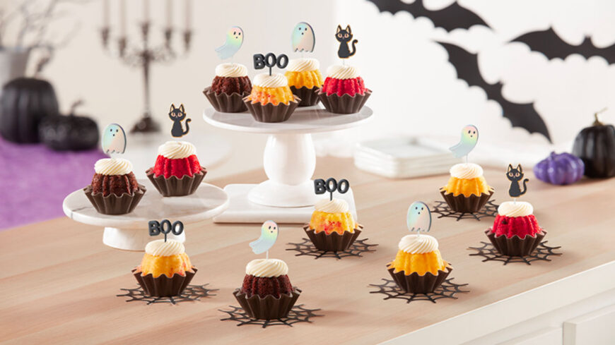 Mini Bundt cakes in chocolate, red velvet, and golden flavors topped with white frosting and Halloween decorations, including ghost and black cat toppers, displayed on white cake stands and a wooden table with bat and pumpkin decorations.