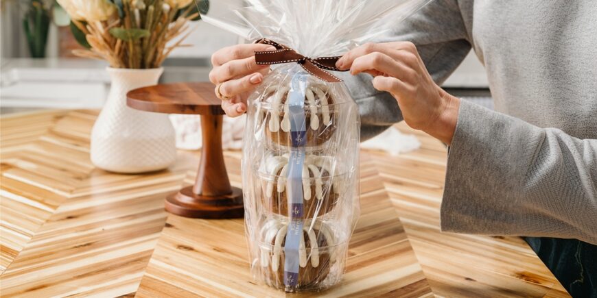 A person ties a brown ribbon around a cellophane-wrapped gift tower of three chocolate Bundt cakes drizzled with white icing.
