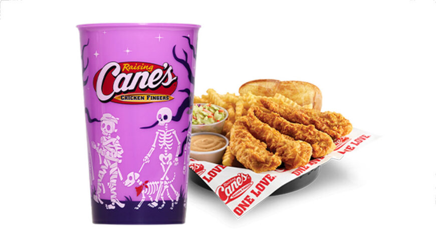 A purple Raising Cane’s Halloween cup decorated with skeletons and spooky designs sits beside a meal of crispy chicken fingers, crinkle-cut fries, Texas toast, coleslaw, and dipping sauce.