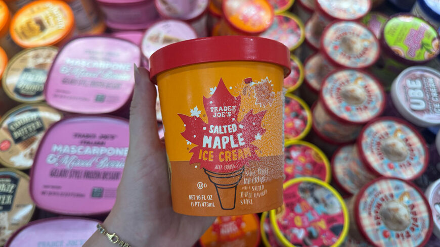 Hand holding a Trader Joe’s Salted Maple Ice Cream container in front of a display of assorted ice cream tubs.