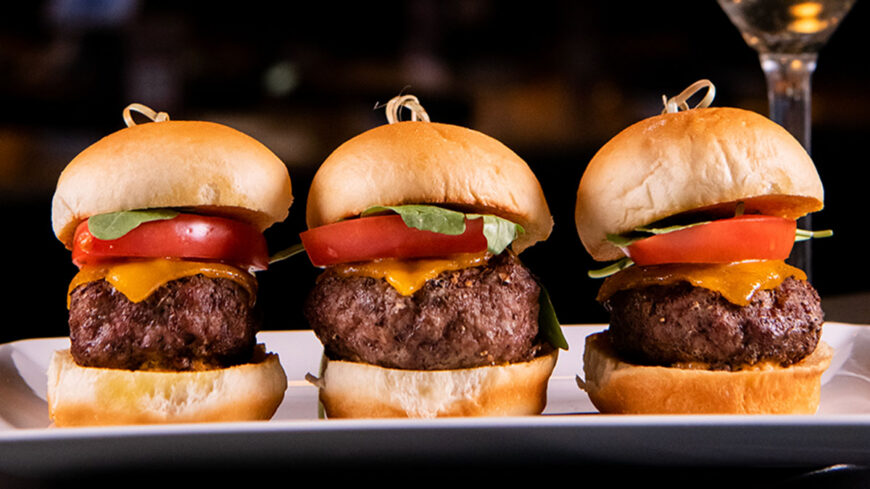 Three grilled beef burger sliders topped with melted cheddar cheese, fresh tomato slices, and lettuce on toasted golden buns, arranged side by side on a white serving platter against a dark background.