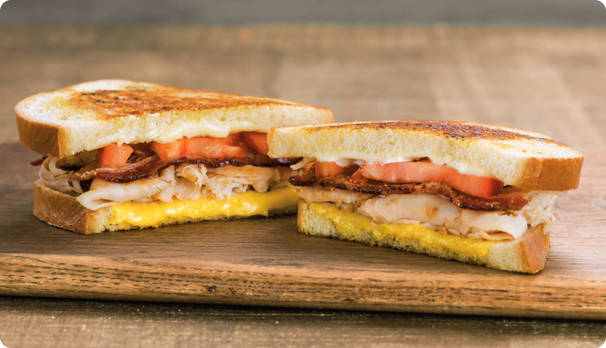 Grilled bacon and turkey sandwich with melted cheddar cheese, tomato and mayonnaise on toasted bread, cut in half and served on a wooden board.