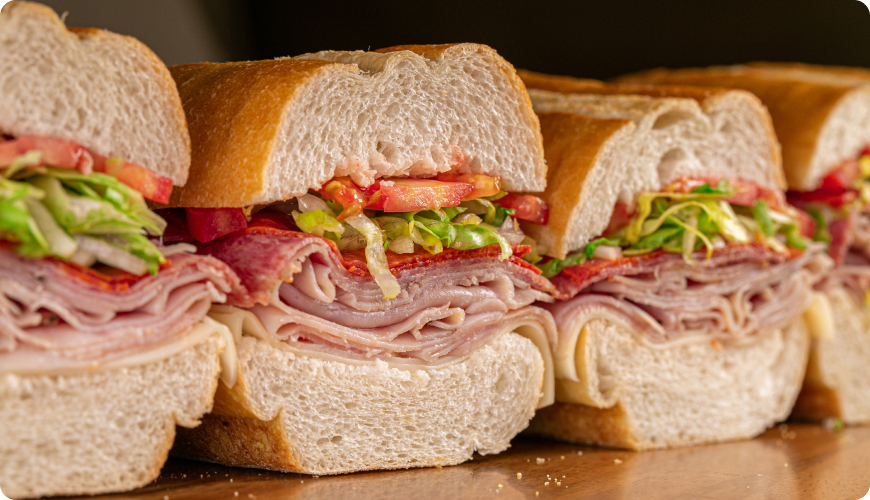 Close-up of a classic deli sub sandwich made with sliced ham, salami, and other deli meats, topped with lettuce, tomatoes, and onions on fresh white bread. Several sandwiches are arranged side by side.