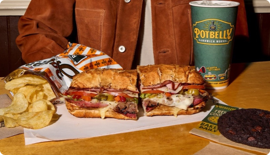 Toasted sandwich cut in half, filled with sliced deli meats, melted cheese, tomatoes, and peppers, served with potato chips, a chocolate cookie, and a Potbelly-branded drink.