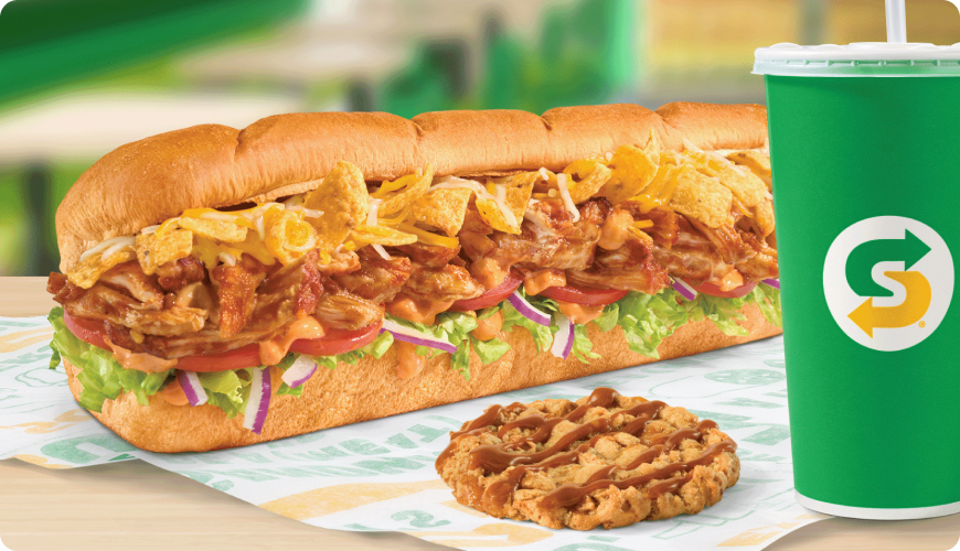 A Subway Chicken Enchilada sub filled with seasoned chicken, melted cheese, tomatoes, lettuce, red onions, and sauce, served alongside a chocolate chip cookie and a large green Subway drink cup.