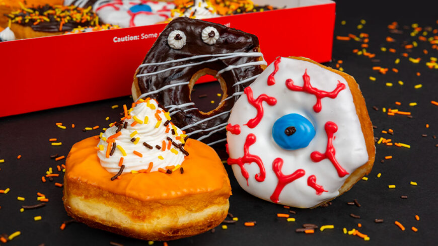Three Halloween-themed donuts featuring an orange glaze with whipped frosting and sprinkles, a chocolate-glazed monster donut with white icing details, and a white-glazed eyeball donut with red icing veins and a blue center, displayed against a dark background with scattered Halloween sprinkles.
