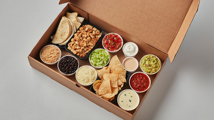 Open cardboard takeout box filled with tortillas, seasoned grilled chicken, rice, black beans, shredded lettuce and cheese, tortilla chips, guacamole, salsa, sour cream, queso, and pico de gallo.