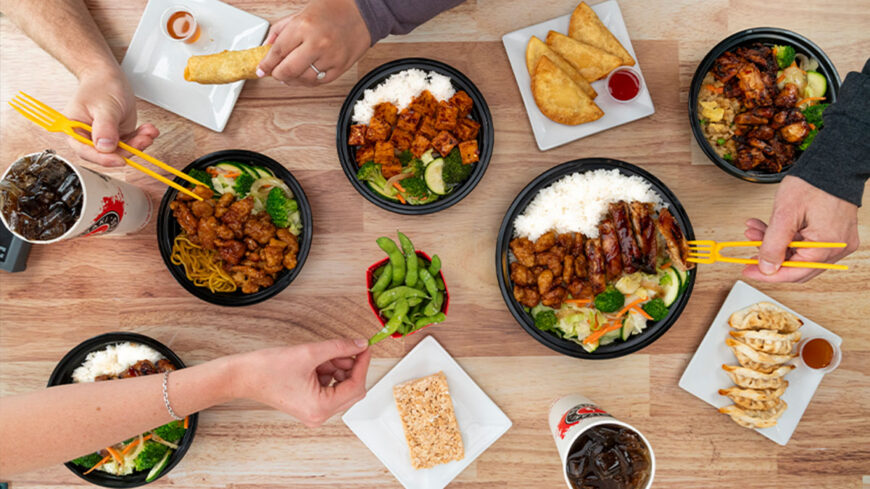 Overhead view of a table filled with teriyaki meals, including rice bowls with grilled chicken, beef, noodles, and vegetables, alongside dumplings, spring rolls, edamame, dipping sauces, and iced drinks as people share the food.