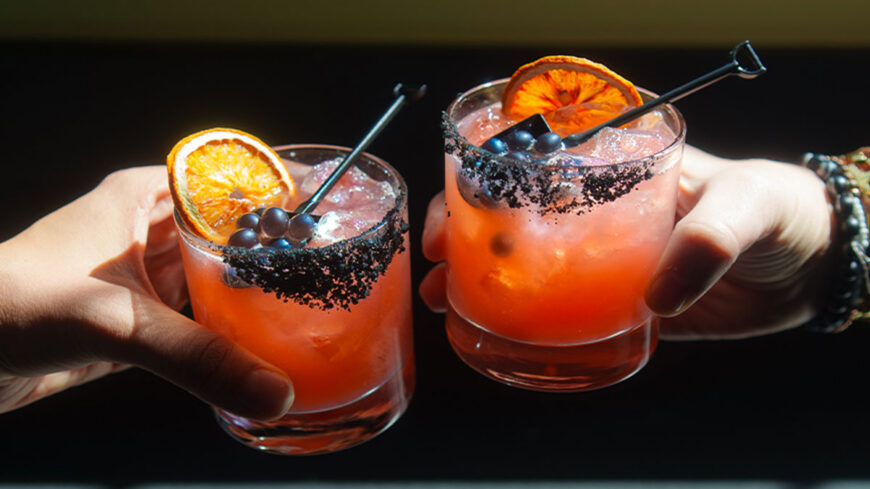 Two hands hold orange-pink Grave Digger margaritas in short glasses with black salt rims, ice, dried orange garnishes, and dark berries, raised together against a dark background.