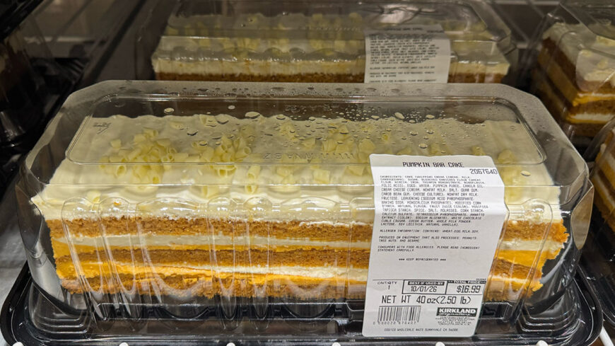 A packaged rectangular pumpkin bar cake with multiple layers of cake, cream filling, and pumpkin-colored frosting, labeled “Pumpkin Bar Cake."